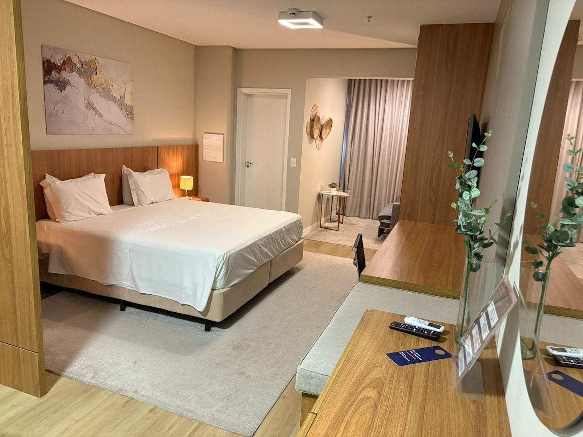 A spacious room is equipped with two beds made with crisp white linens, complemented by warm wooden accents. A soft rug covers the floor, and a sleek desk sits adjacent to a modern TV. Natural light filters through sheer curtains, creating a calm ambiance.