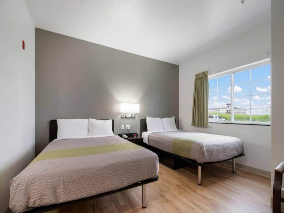 A well-lit guest room features two spacious beds with light-colored bedding. A nightstand is positioned between the beds, equipped with bedside lamps. Natural light enters through a window with sheer curtains, illuminating the neutral-toned walls and wooden flooring.