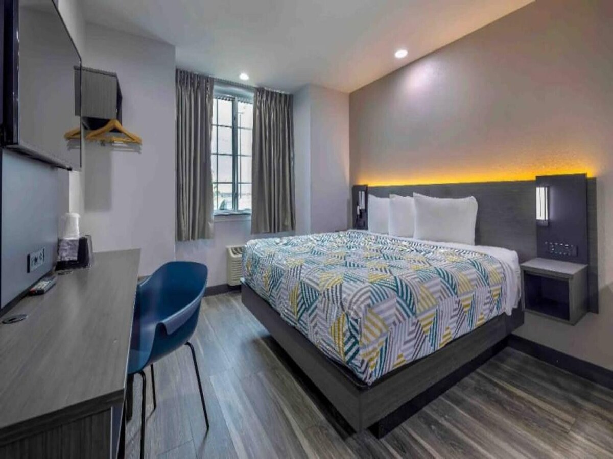 A spacious room features a king-sized bed with a patterned duvet and two white pillows. Soft lighting is integrated into the wall behind the bed. A window with light drapes allows natural light, and a desk with a chair is included.
