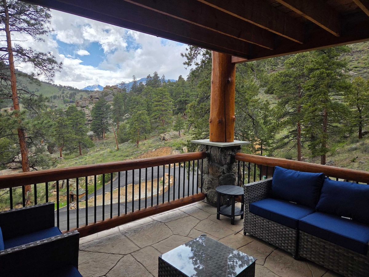A spacious outdoor deck is furnished with a seating area featuring navy blue cushions and a glass-top table. The surrounding views include lush green trees and distant mountains, with a stone wall adding texture to the space. The atmosphere appears serene and inviting.