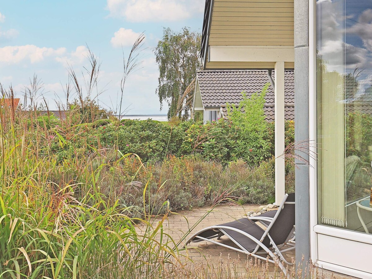 A quiet outdoor space is highlighted by a pair of reclining chairs positioned on a patio. Lush greenery and flowering plants create a natural border, while a glimpse of neighboring houses is visible in the background, set against a clear sky.