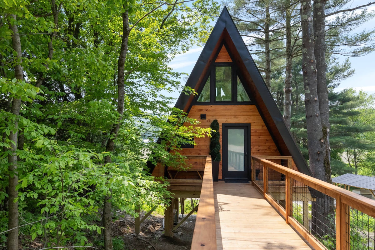 The A-frame treehouse entrance is framed by lush greenery, showcasing a blend of wood and modern finishes. A wooden deck leads to the front door, surrounded by tall trees, while large windows reflect natural light and provide a glimpse of the inviting interior.