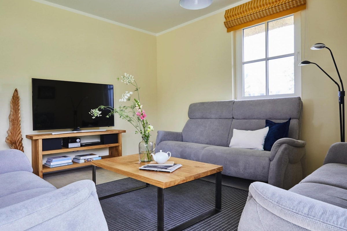 A living room features a comfortable seating area with a grey sofa and armchairs. A wooden coffee table is centered, adorned with a decorative arrangement of flowers and a tea set. A large television is positioned on a wooden stand, and natural light enters through a nearby window.