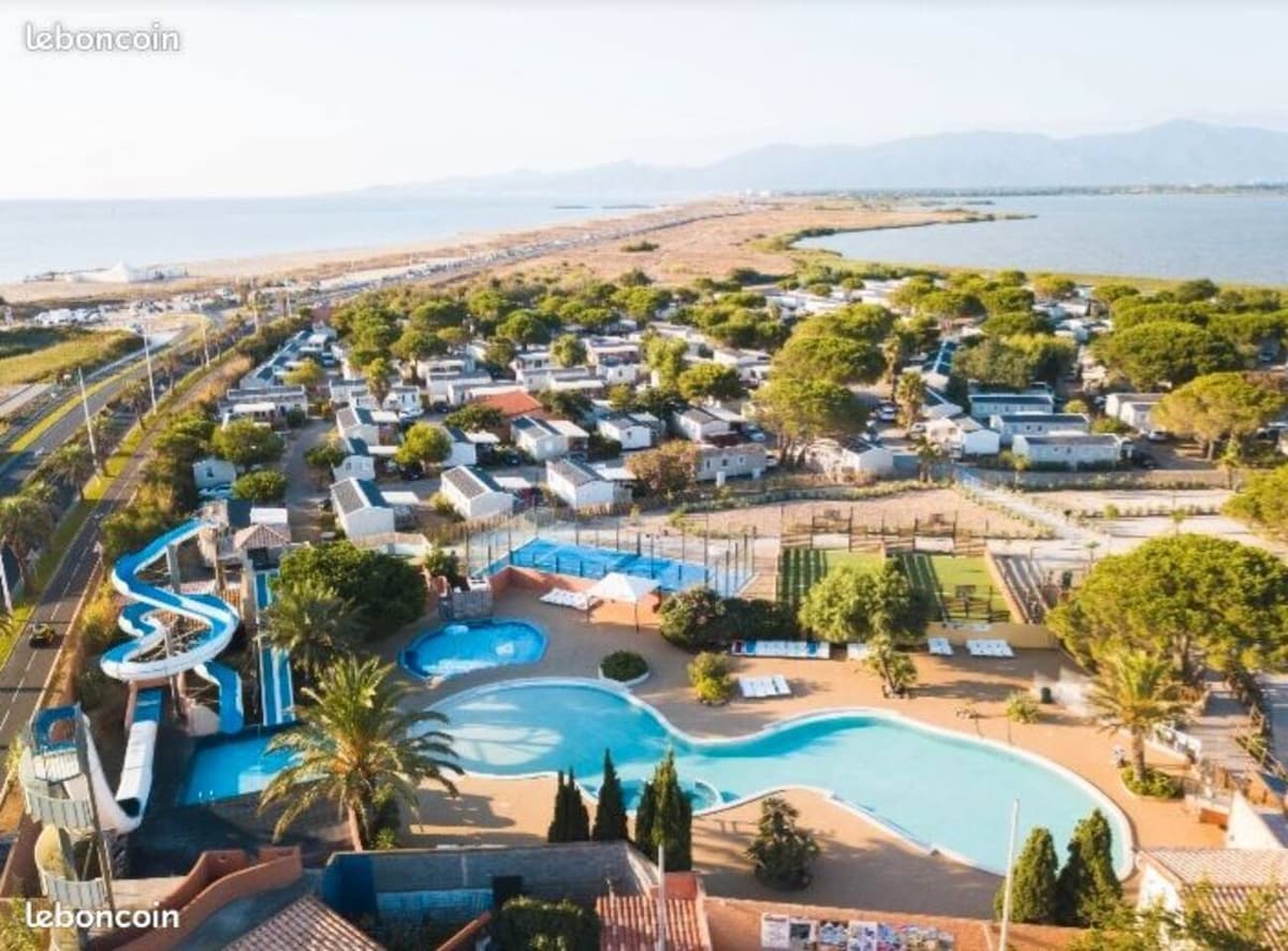 Aerial view of the campsite showcasing a large swimming pool with slides, surrounded by palm trees. Various mobile homes are positioned throughout the site, alongside recreational areas and sports fields. The beach and mountains are visible in the background, emphasizing the natural setting.