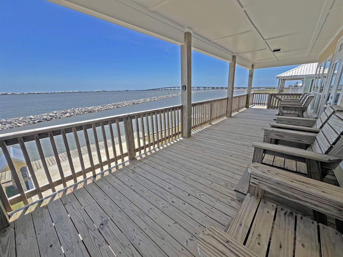 A spacious covered deck is provided with light wooden flooring and numerous Adirondack chairs facing the water. The view includes a serene bay and a distant bridge, with bright blue skies overhead, creating a relaxed outdoor atmosphere.