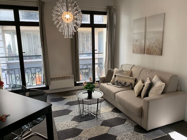 A living room features large windows allowing natural light to fill the space. A light-colored sofa with decorative cushions is arranged around a circular coffee table. A unique light fixture hangs above, complementing the patterned area rug beneath. Two wall art pieces are displayed nearby.