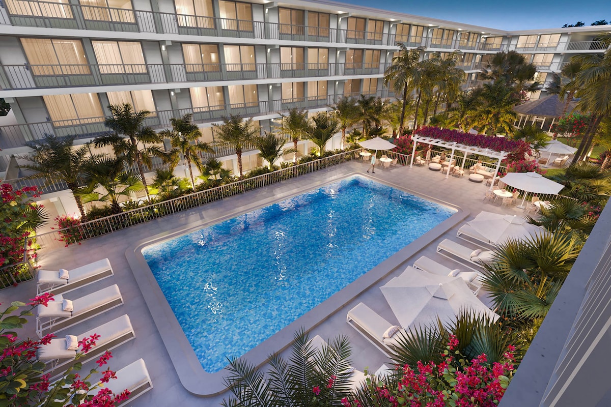 A resort-style pool is surrounded by lush greenery and colorful flowers. Lounge chairs are placed around the pool, offering a relaxing space. The area is softly illuminated, enhancing the inviting atmosphere in the evening light.
