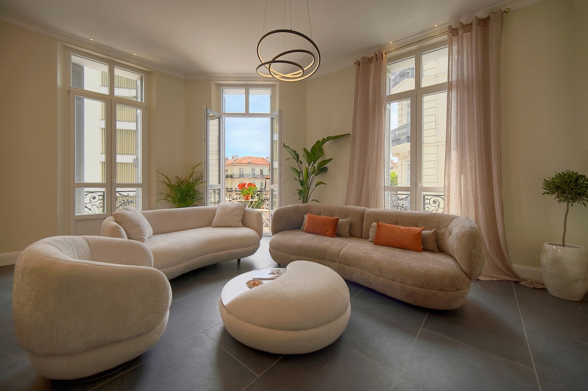 A bright living room features a modern curved sofa and chair set, complemented by a circular coffee table. Large windows allow natural light to fill the space, while delicate curtains frame the view. Potted plants add a touch of greenery to the elegant atmosphere.
