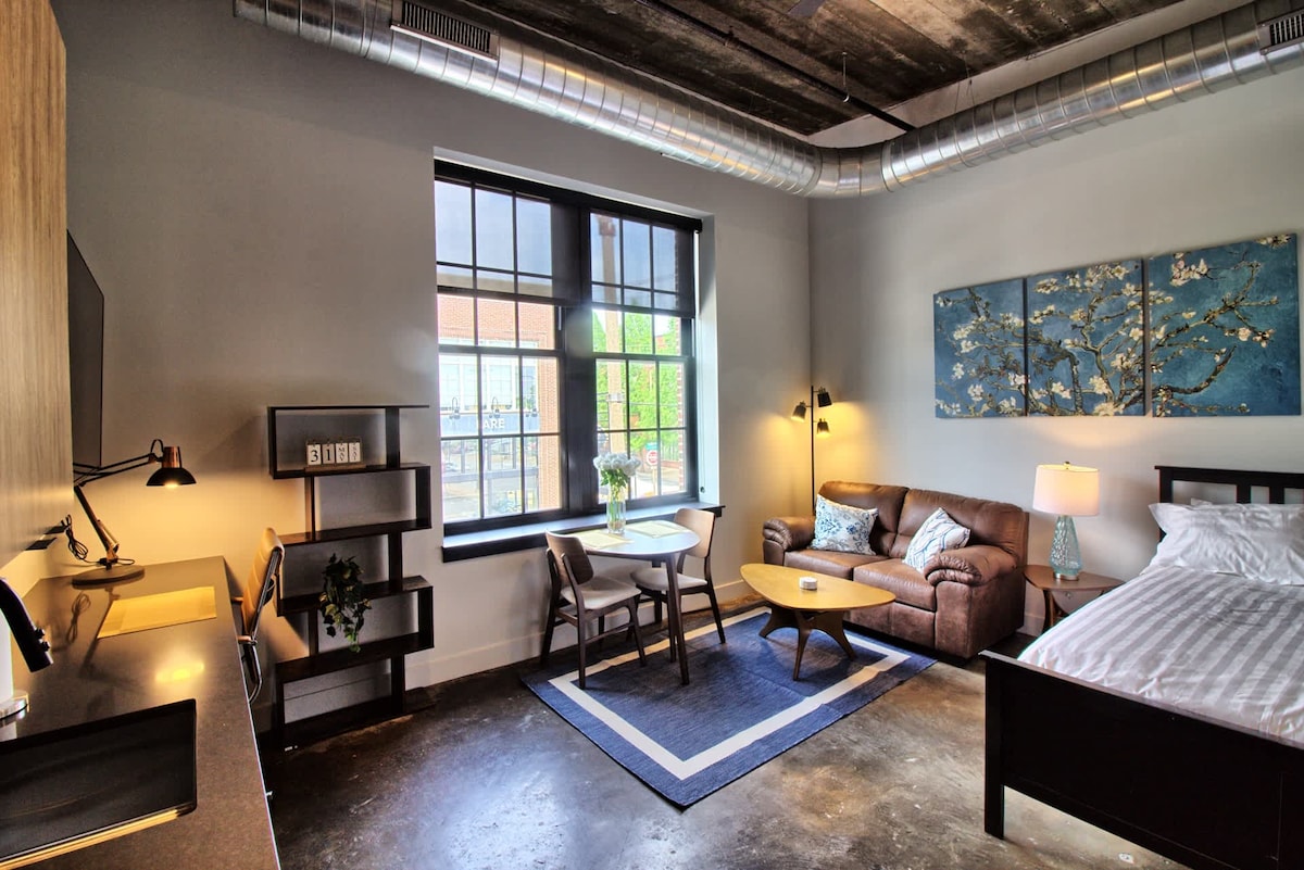 A minimalist studio features high ceilings and industrial-style elements. A comfortable seating area is arranged near a window, allowing natural light to enter. A small dining table and stylish decor complement the space, which includes a spacious bed and modern furnishings.
