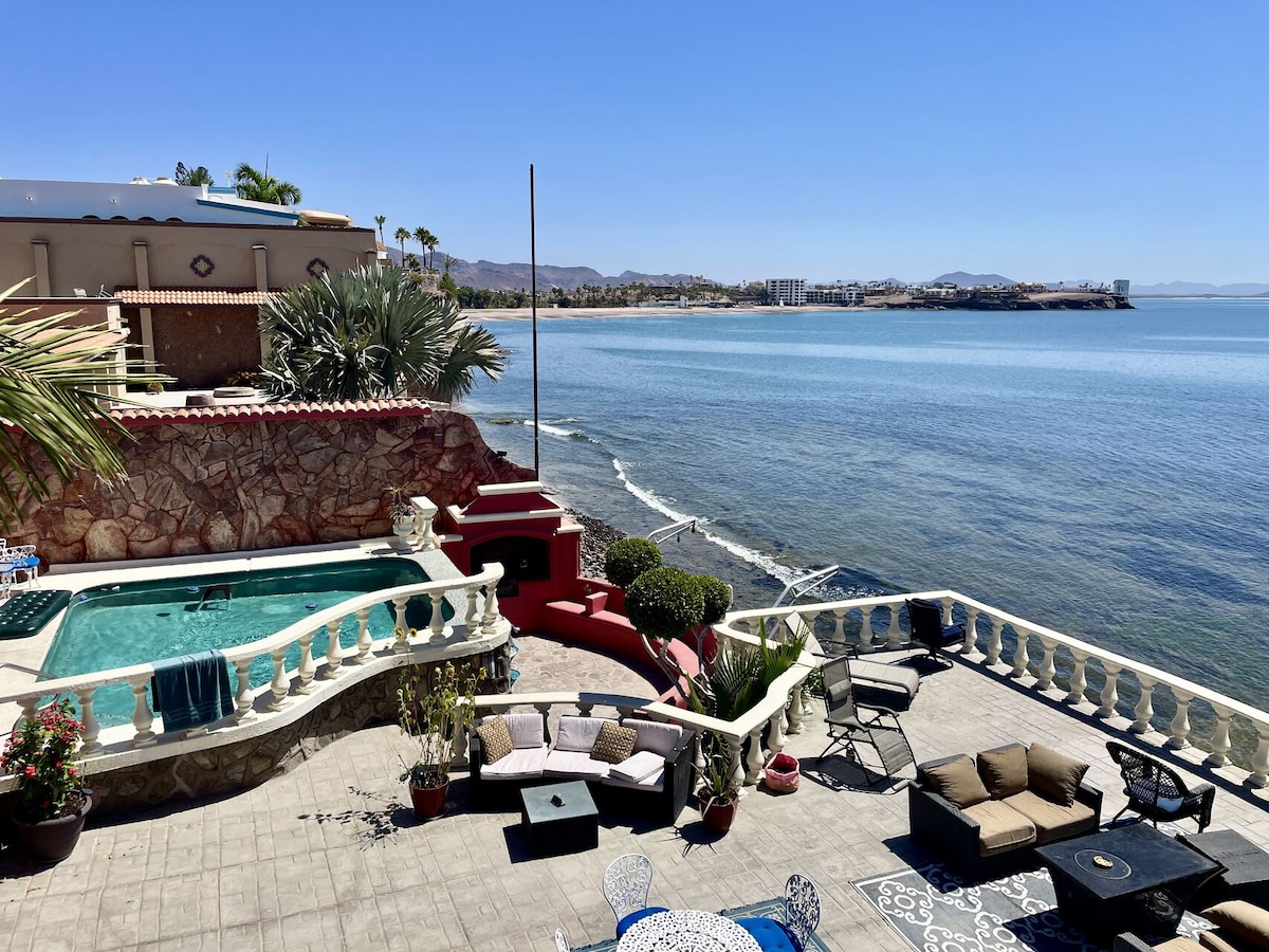 An outdoor area features a private swimming pool surrounded by potted plants and comfortable seating options. The view overlooks the tranquil water and coastline, with distant buildings visible along the shore. Palm trees add a touch of greenery to the serene environment.