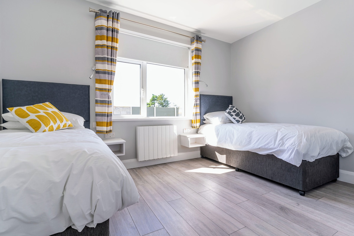 The room features two twin beds with white bedding and decorative pillows. Large windows allow natural light to fill the space, framed by grey and yellow patterned curtains. Light wooden flooring enhances the bright and airy atmosphere.