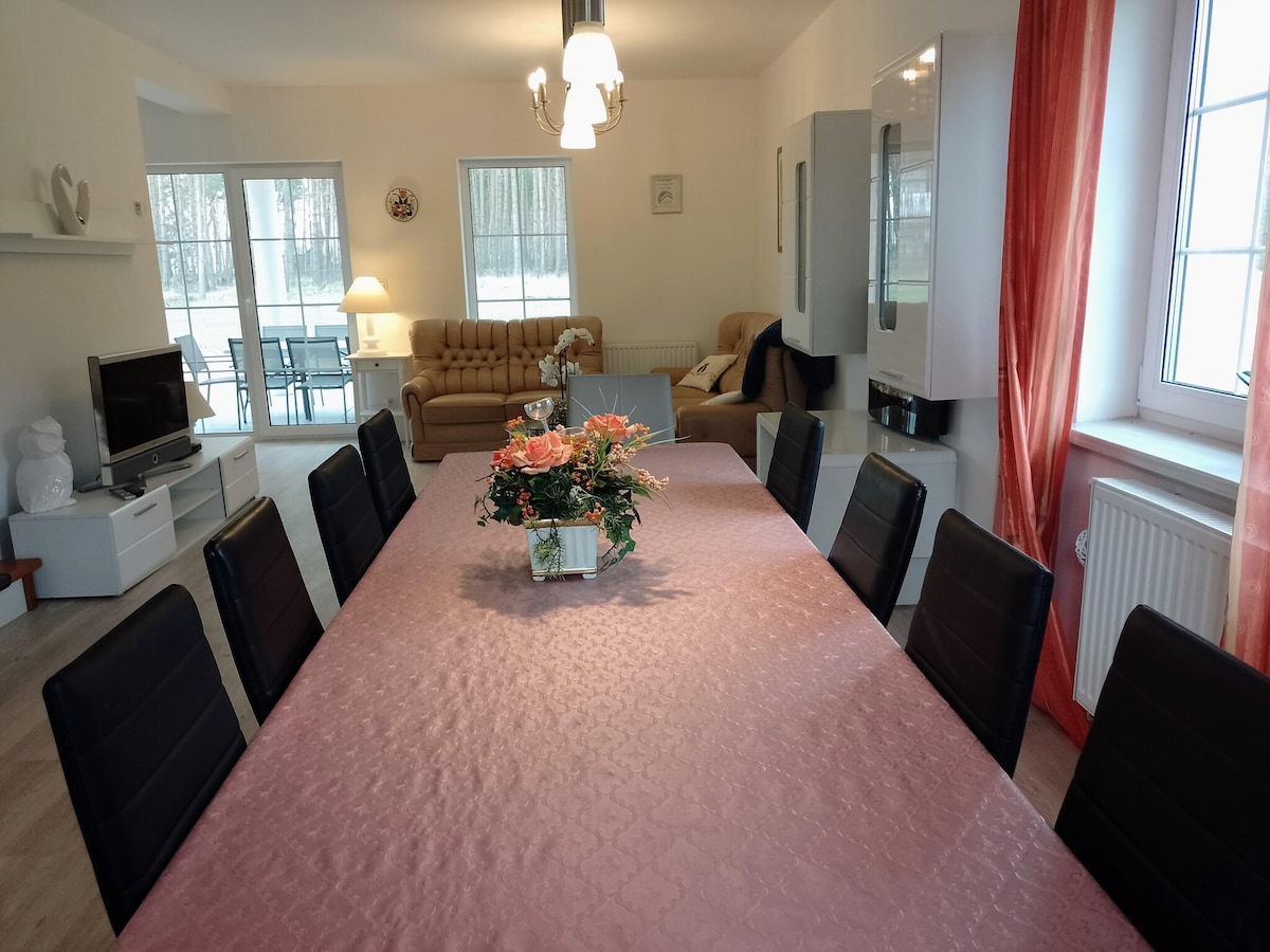 A spacious dining area features a long table set with a light pink tablecloth, surrounded by black chairs. A floral centerpiece is placed prominently on the table. In the background, a comfortable seating area is seen, with large windows allowing natural light to illuminate the room.
