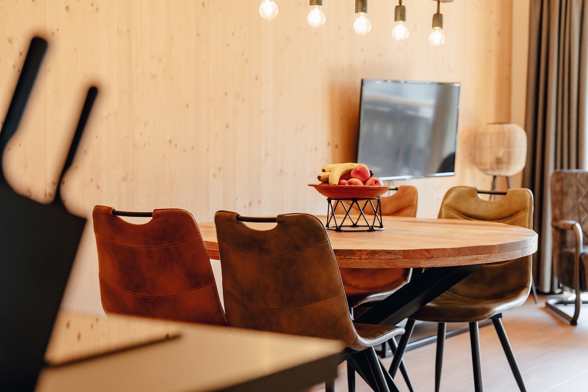 A round wooden dining table is surrounded by comfortably cushioned chairs in varying shades. A bowl of fresh fruit is displayed at the center. A wall-mounted Smart TV and stylish lighting fixtures complement the cozy ambiance within the dining area.