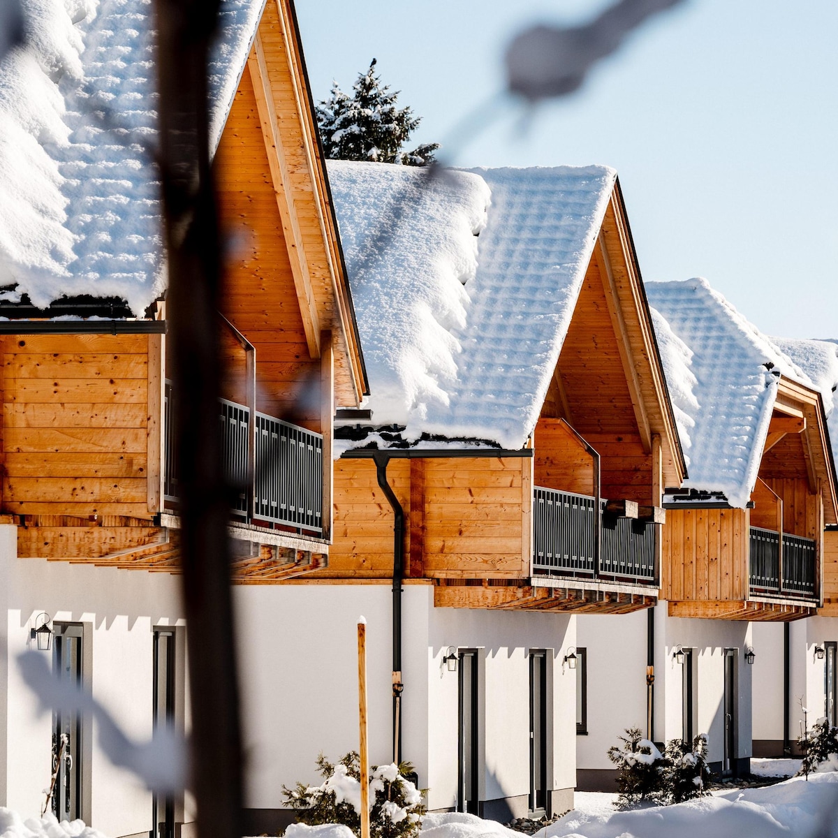 Architecturally designed chalets are seen, with sloped roofs covered in a layer of fresh snow, creating a cozy winter atmosphere. Balconies with wooden railings are featured prominently, providing outdoor space. The facades are painted in neutral tones, contrasting gently with the white snow.