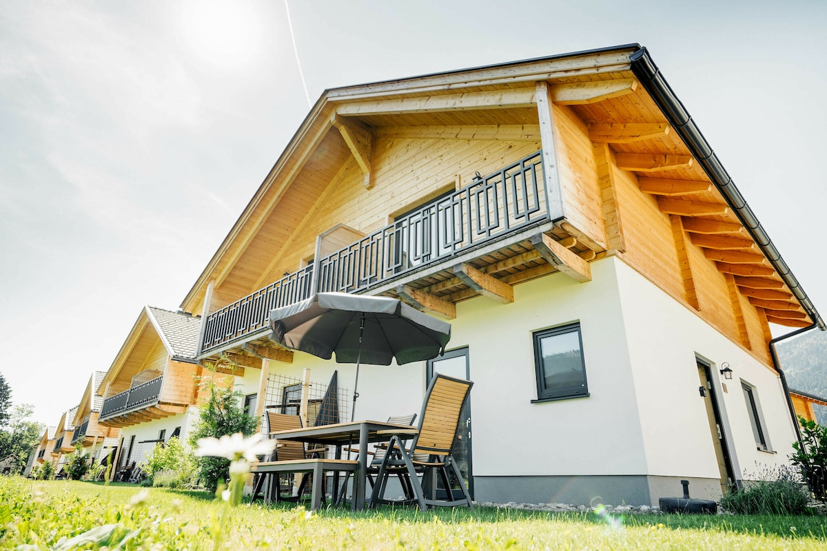 A luxurious chalet features a wooden exterior, with a sloped roof and a balcony adorned with a black railing. Outdoor seating with a table and chairs is present on a grassy area, providing a welcoming space for relaxation.