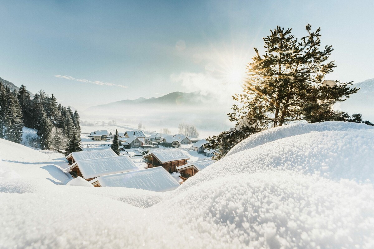 A serene winter landscape is depicted, showcasing snow-covered rooftops nestled among rolling hills and evergreen trees. The sun rises gently over the mountains in the distance, casting a warm glow on the snowy scene.