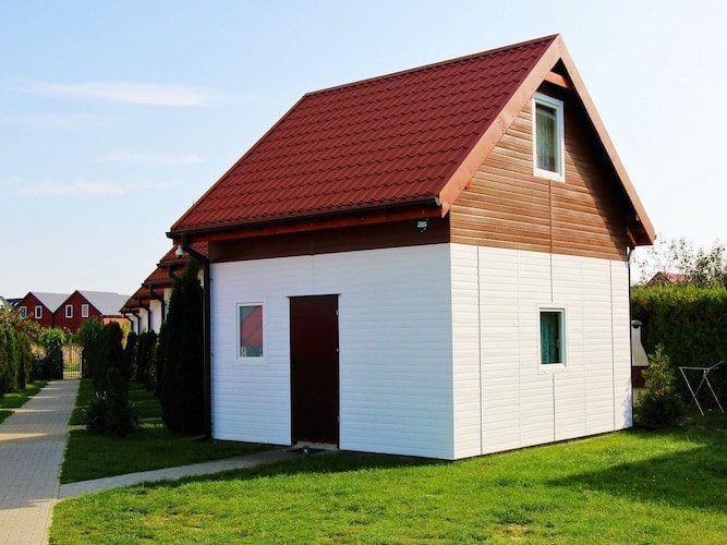 A two-storey holiday home is presented, featuring a combination of white and wooden exterior walls. A pitched roof with red tiles complements the rustic design. The entrance door and windows are visible, surrounded by well-maintained grass, creating a welcoming appearance in the quiet resort.