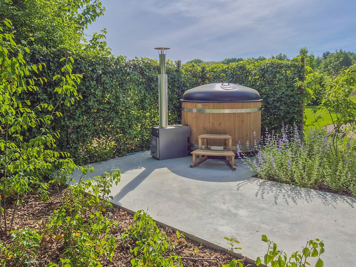 A private hot tub is set on a concrete pad surrounded by lush greenery and flowering plants. A wooden bench is positioned nearby, and a chimney stands beside the hot tub, providing warmth and comfort for a relaxing outdoor experience.
