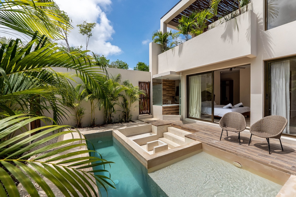 A private pool is highlighted within a modern villa, surrounded by lush greenery. Two lounge chairs are positioned beside the pool, inviting relaxation. Large glass doors offer a view into the living area, where natural light fills the space.