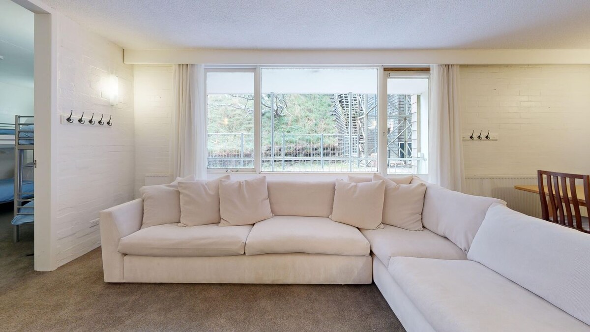 A spacious lounge area features a soft, light-colored sectional sofa arranged for comfort. Large windows allow natural light to fill the space, with views of the outdoors visible. The room includes a dining table, blending functionality with a relaxed atmosphere.