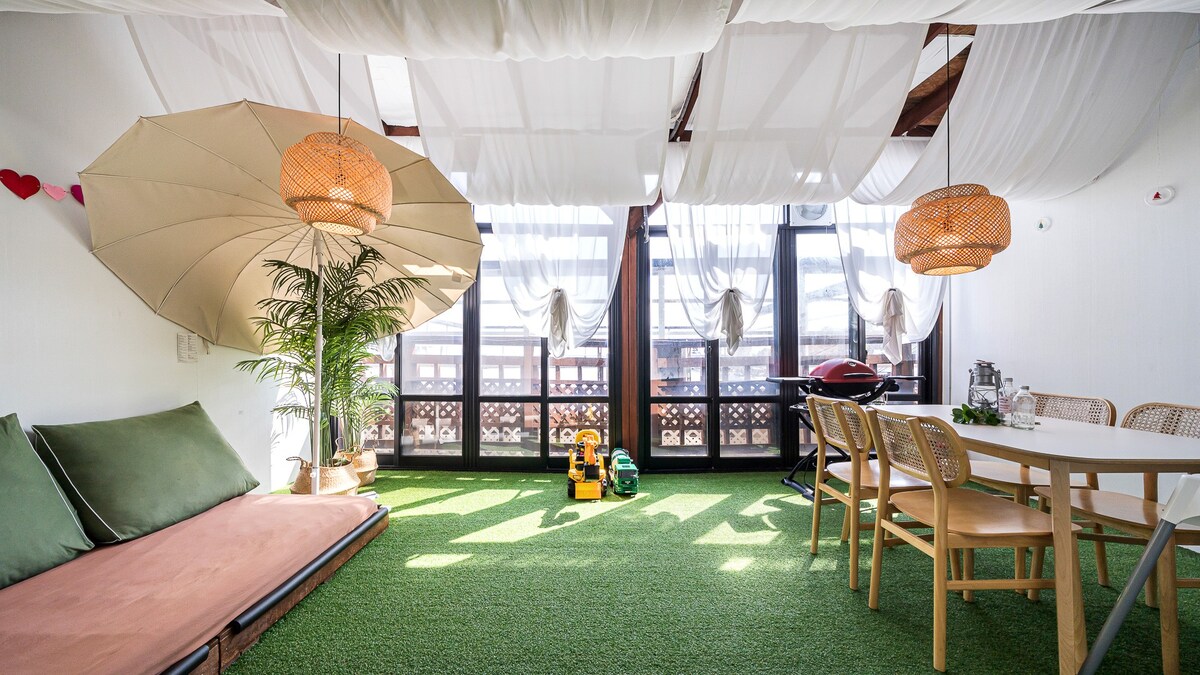 A bright and airy space features green artificial grass flooring, illuminated by natural light. Two large woven pendant lights hang from the ceiling, casting gentle shadows. A cozy seating area with a soft sofa and two large umbrellas adds charm. A dining table awaits, complemented by simple wooden chairs.