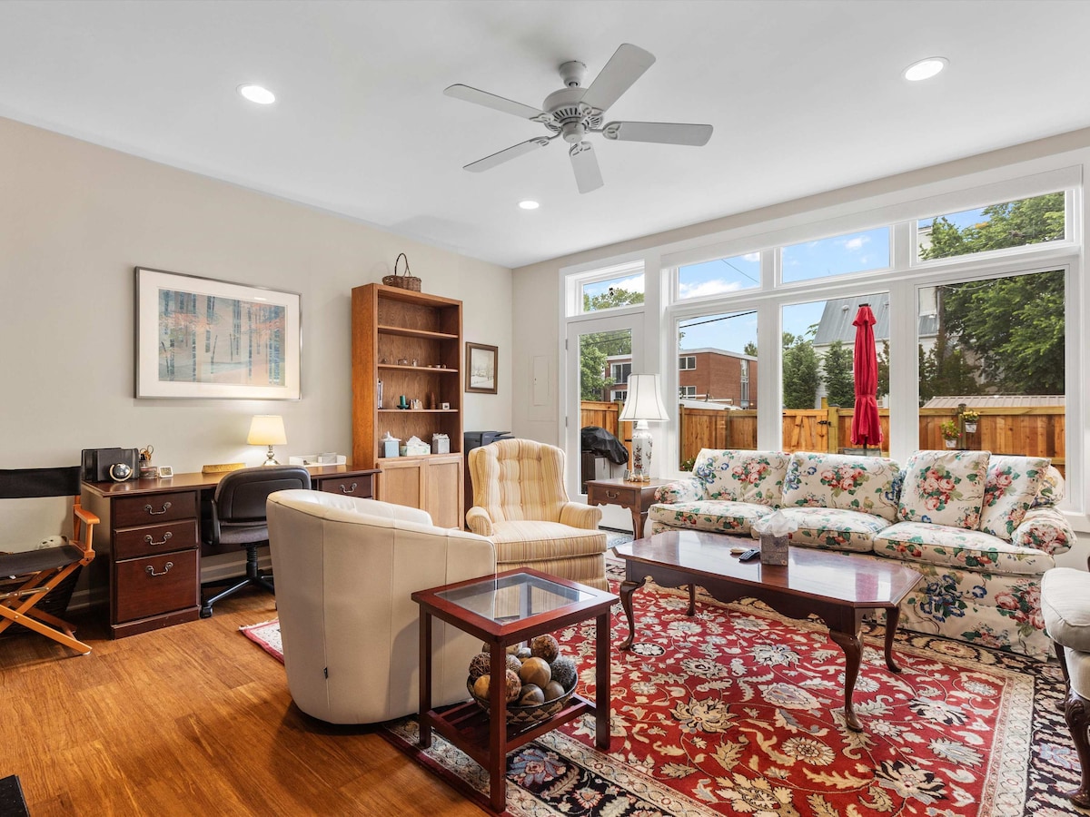 A spacious living room is highlighted by large windows that allow natural light to fill the space. Comfortable seating includes a floral-patterned sofa and a single armchair, complemented by a decorative area rug. A wooden desk and shelf provide additional functional elements.