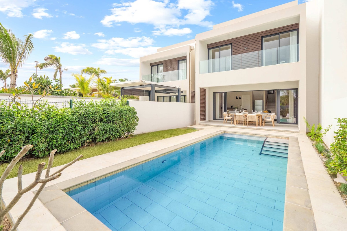 The exterior view of Villa Halona features a modern design with large glass doors opening to the outdoor area. A large swimming pool, bordered by light-colored stone tiles, reflects the sky. Lush greenery and palm trees enhance the serene atmosphere, while a covered terrace accommodates outdoor dining.