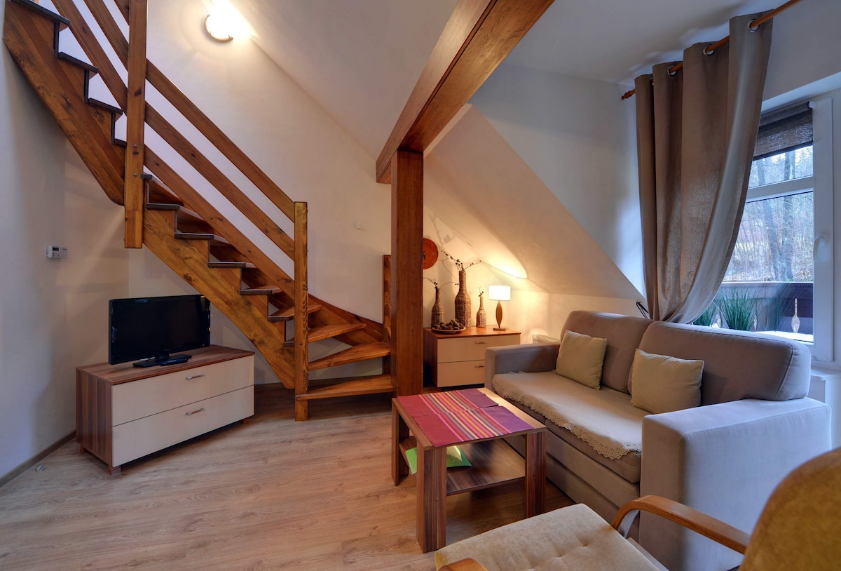 ห้องนั่งเล่นทันสมัยพร้อมโซฟา ทีวีจอแบน หน้าต่างบานใหญ่ และบันไดที่มองเห็นได้ซึ่งนำไปสู่ชั้นบน