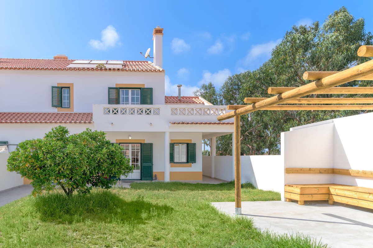 The villa's exterior features a well-maintained garden with lush grass and surrounding trees. A wooden pergola is present on a paved patio area, providing shaded seating. The property showcases traditional architectural elements, including green shutters and terracotta roofing under a clear blue sky.