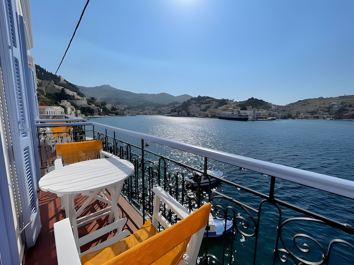A serene balcony overlooks a calm sea with gentle waves reflecting sunlight. White outdoor furniture is arranged neatly, complemented by bright orange cushions. The distant town of Symi and its rolling hills are visible, creating a tranquil coastal atmosphere.