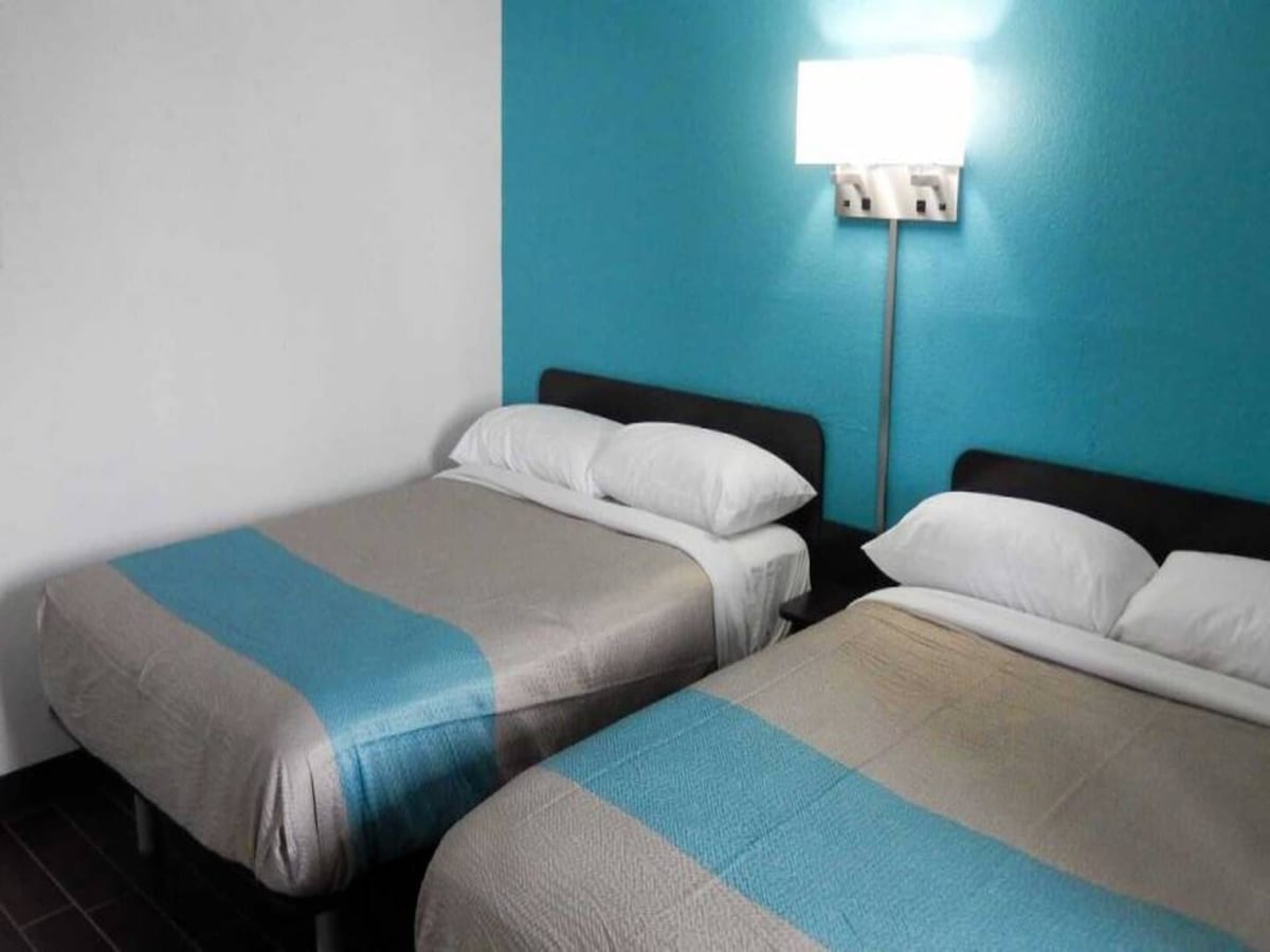 A comfortable bedroom is shown with two queen beds covered in neutral-toned linens featuring a light blue stripe. A turquoise accent wall adds a pop of color. Soft lighting is provided by a modern wall-mounted lamp.