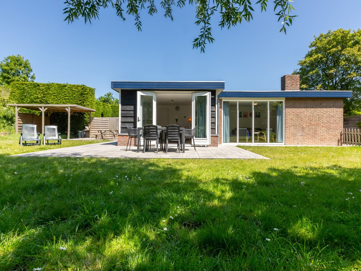 The holiday home features a spacious patio area surrounded by a lush green garden. Outdoor dining furniture accommodates several guests, while two sun loungers invite relaxation. Large glass doors lead into the interior, creating a seamless connection between indoor and outdoor spaces.