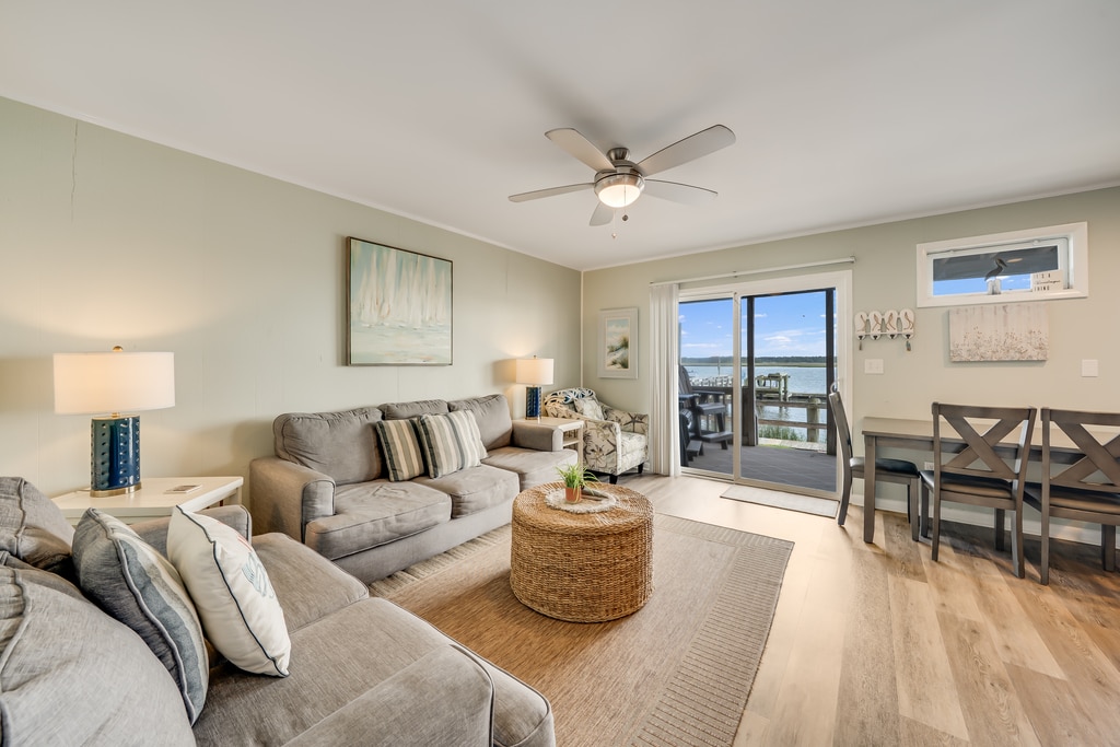 A bright and inviting living area features a comfortable seating arrangement with two light gray sofas and a round coffee table. Natural light filters in through sliding glass doors that lead to a balcony, with a dining table and four chairs positioned nearby.