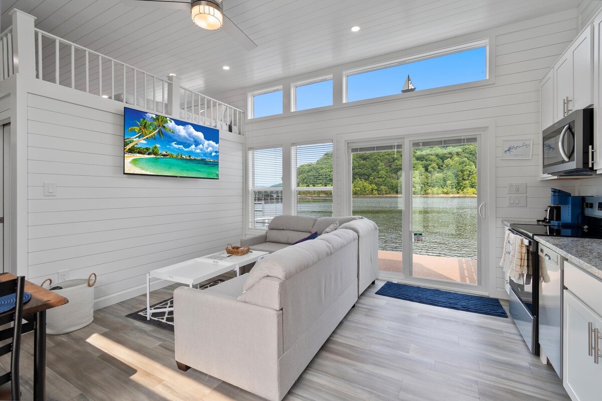 A bright living area features large windows offering expansive lake views. A light-colored sectional sofa is positioned in front of a mounted television. The modern kitchen is visible, equipped with stainless steel appliances, and a dining table is nearby, all reflecting a clean and contemporary design.