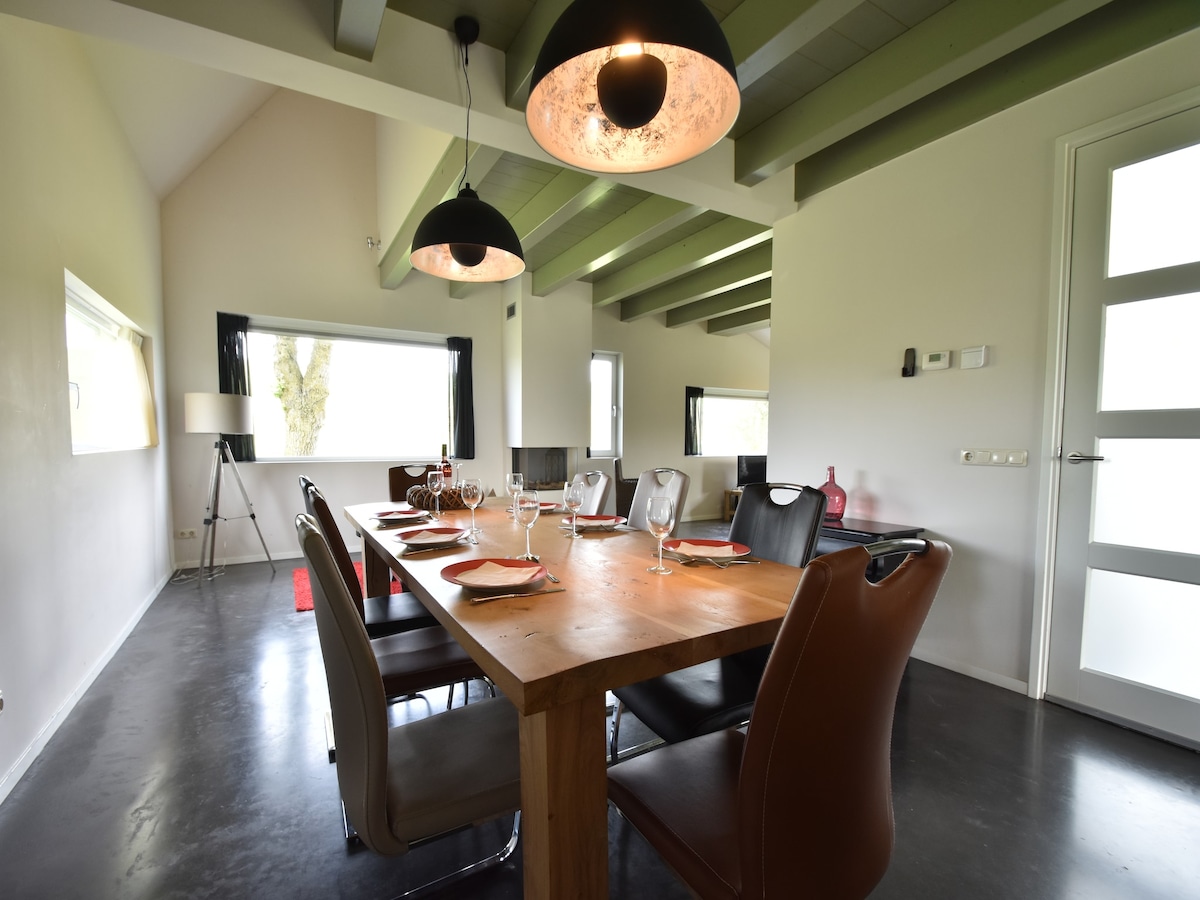 A spacious dining area is highlighted by a long wooden table set for eight, with elegant tableware arranged thoughtfully. Stylish seating options surround the table, complemented by large windows allowing natural light to brighten the room. Distinctive pendant lights hang from the ceiling, creating a relaxed ambiance.