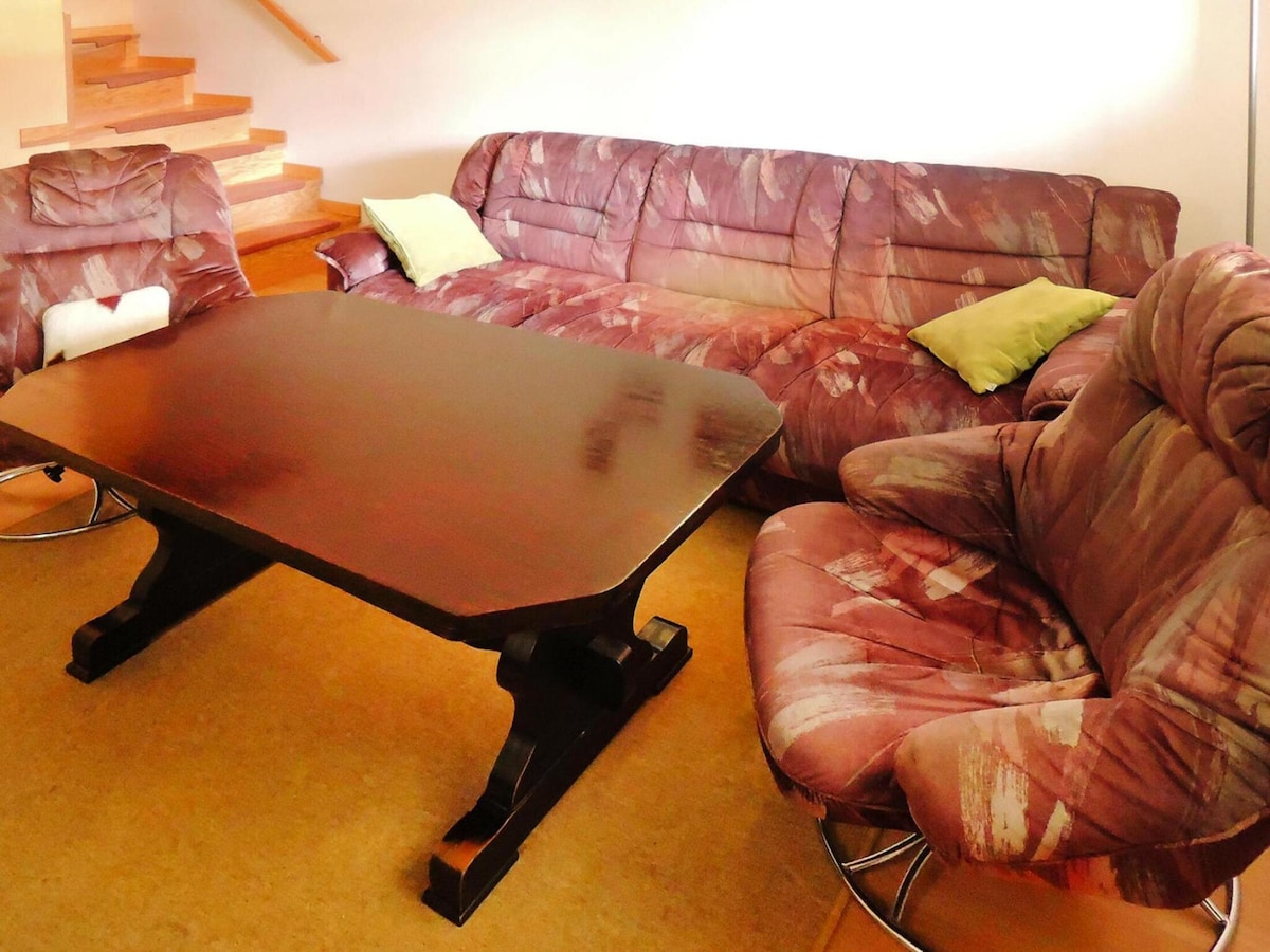 A living area features a spacious dark wood table at the center, surrounded by a plush couch and two large armchairs. The seating is upholstered in a mix of muted colors, complemented by bright cushions. Wooden stairs lead to an upper level in the background.