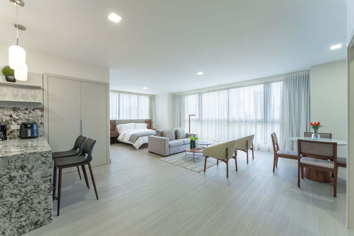 The open living area showcases a modern design, with a comfortable sofa facing a large window draped in sheer curtains. A dining table is positioned near the kitchen, featuring sleek appliances and stylish seating. The bedroom area is seen in the background, characterized by a spacious layout.