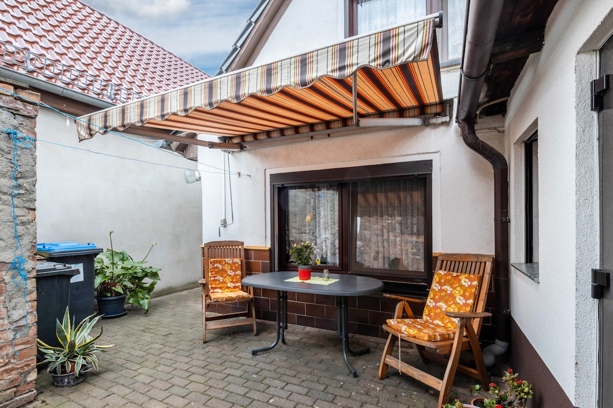 A private outdoor terrace features a striped awning offering shade. Two wooden chairs with cushions are positioned around a small table, creating a cozy seating area. Potted plants and a corner with a trash bin are visible in the background.