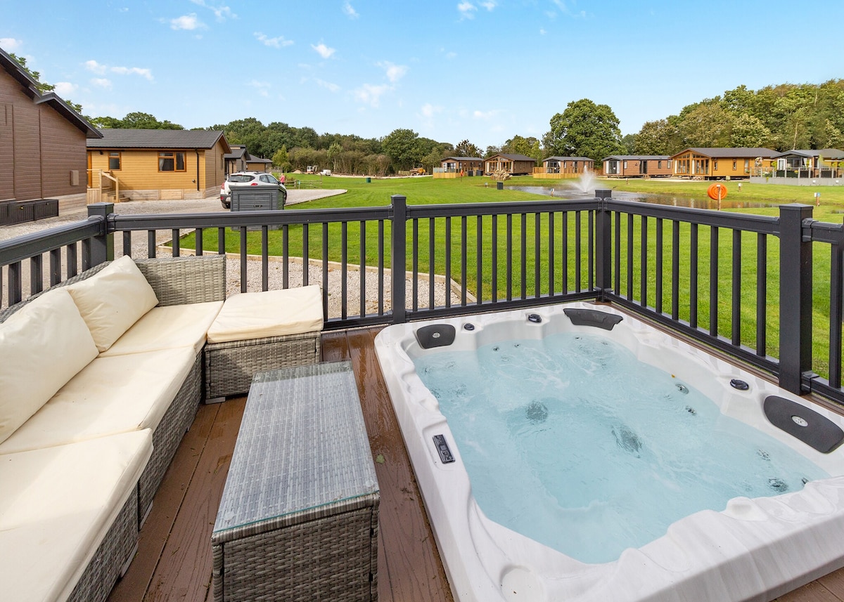 An outdoor deck area features a hot tub and comfortable seating on a stylish couch, surrounded by lush green lawns and several lodge accommodations in the background. The serene environment highlights the tranquil setting of the lodge.