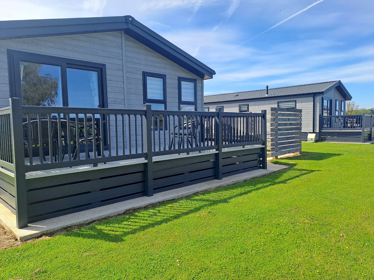 Two modern holiday lodges are depicted, featuring spacious decks with railings. The sleek exterior design is complemented by large windows, offering views into the open living spaces. A well-maintained grassy area surrounds the lodges, enhancing the outdoor appeal.