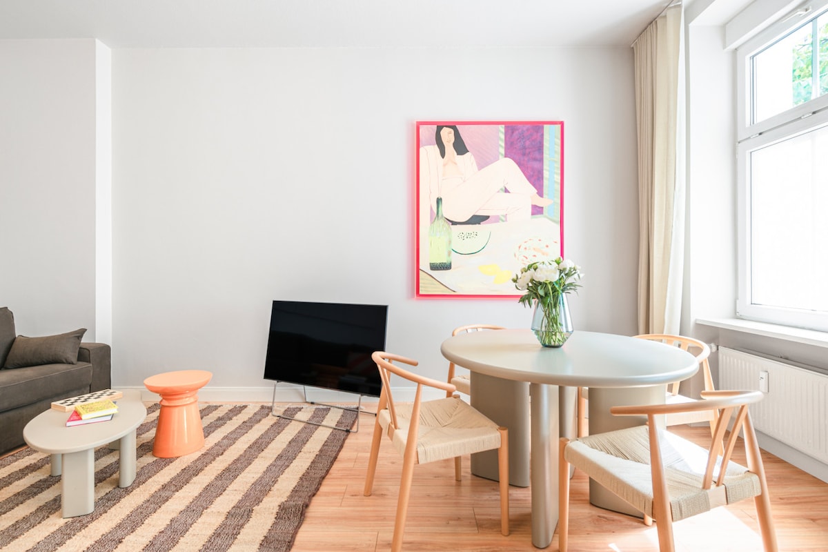 A spacious living area showcases a comfortable grey sofa and a modern coffee table on a patterned rug. A round dining table with wooden chairs is positioned nearby. A large flat-screen TV is visible, and a colorful piece of wall art adds a vibrant touch. Natural light enters through the window.