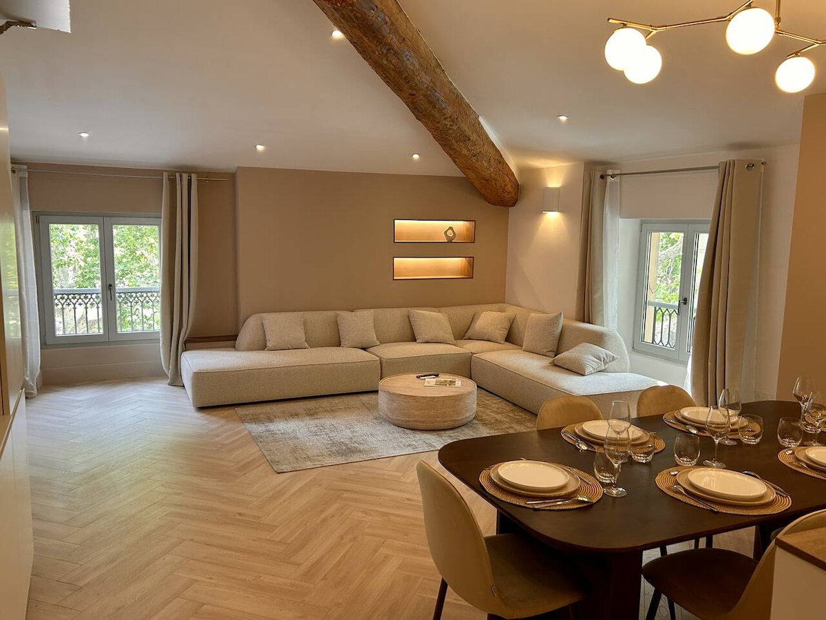 A spacious living area features a large, curved sectional sofa and a round coffee table at its center. Natural light floods the space through large windows with textured curtains. A dining table with elegant table settings complements the inviting atmosphere.