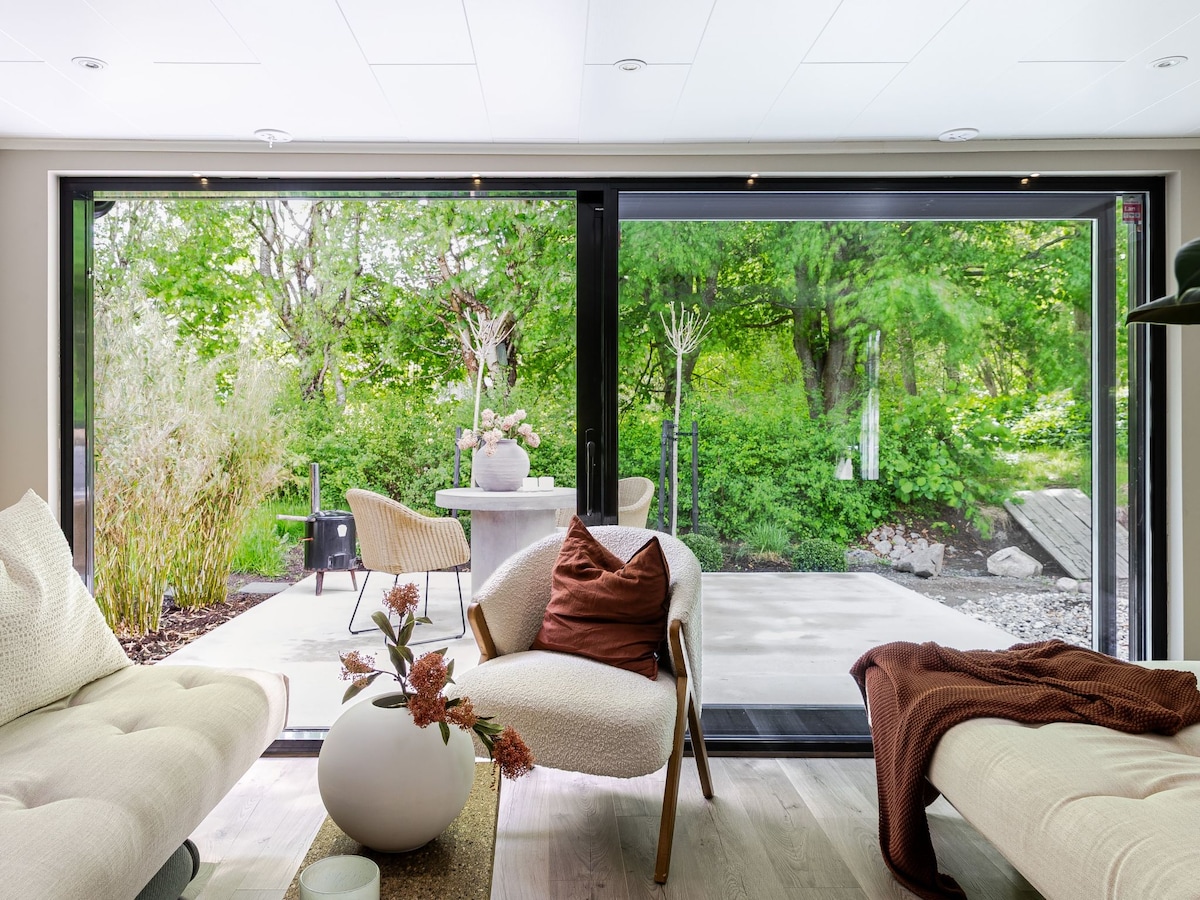 A spacious living area showcases large glass doors that open to a garden and terrace, surrounded by vibrant greenery. Comfortable furnishings include soft armchairs and a decorative table with floral accents, creating a welcoming indoor atmosphere.