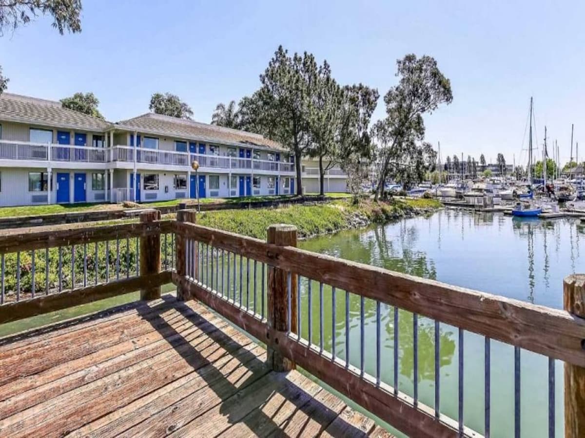 A serene waterfront view showcases a wooden deck extending toward calm waters, reflecting nearby trees and boats. The motel's exterior features a two-story structure with blue doors and windows, surrounded by lush greenery, adding to the peaceful ambiance.