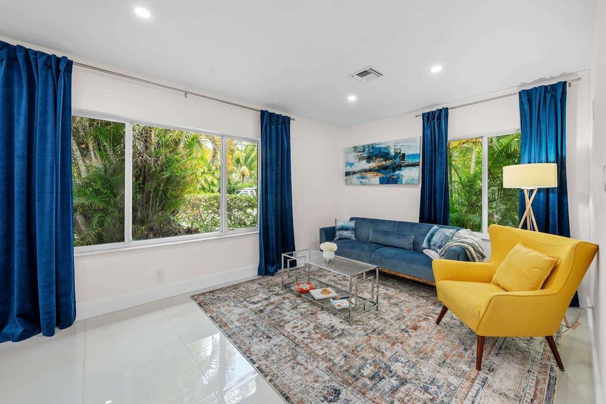 A bright living room is well-lit by natural light through large windows surrounded by tropical greenery. A comfortable blue sofa and a yellow accent chair invite relaxation. A stylish coffee table sits in front of a colorful area rug, enhancing the room's modern decor.