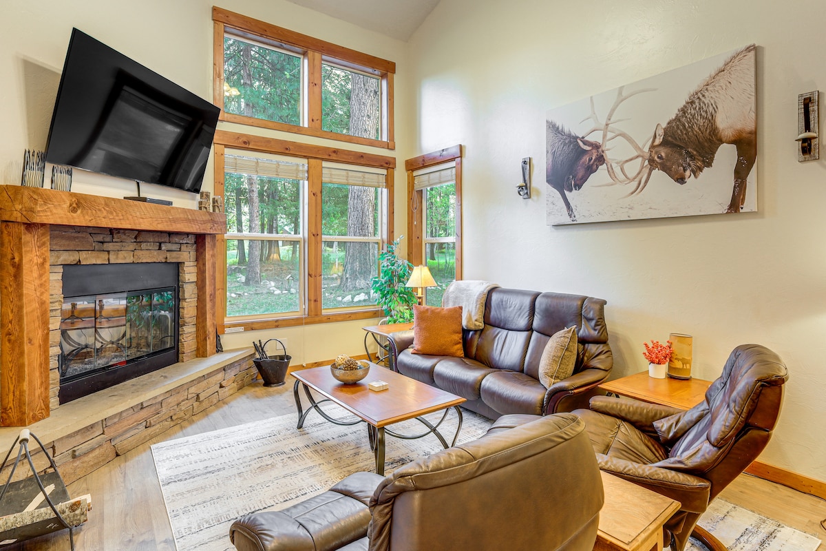 A comfortable living area features a stone fireplace framed by wooden accents, with large windows allowing natural light and views of the outdoors. A mounted flat-screen TV is positioned above the fireplace. Leather sofas and recliners provide ample seating around a wooden coffee table.