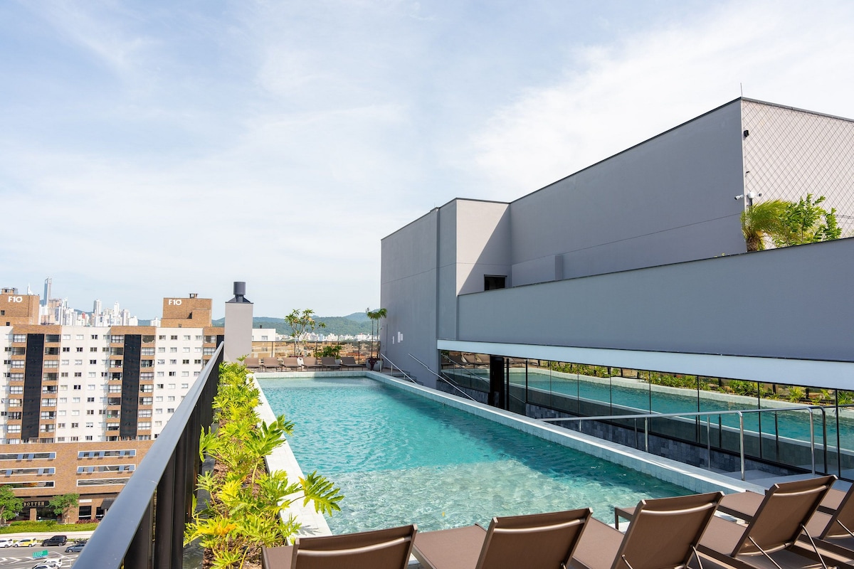 A rooftop area features a long swimming pool with clear water, surrounded by lounge chairs. A modern building and city skyline can be seen in the background, highlighting a welcoming outdoor space for relaxation.