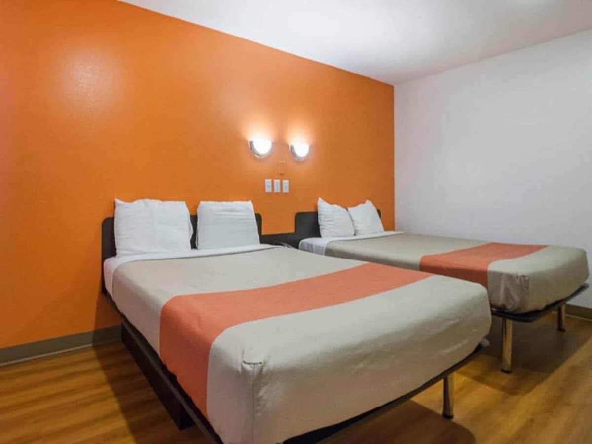A room features two queen beds dressed in light bedding, positioned against an orange accent wall. Two wall-mounted lights provide soft illumination, and the wood flooring creates a warm ambiance. The opposite wall remains neutral and unadorned, contributing to a simple yet functional space.