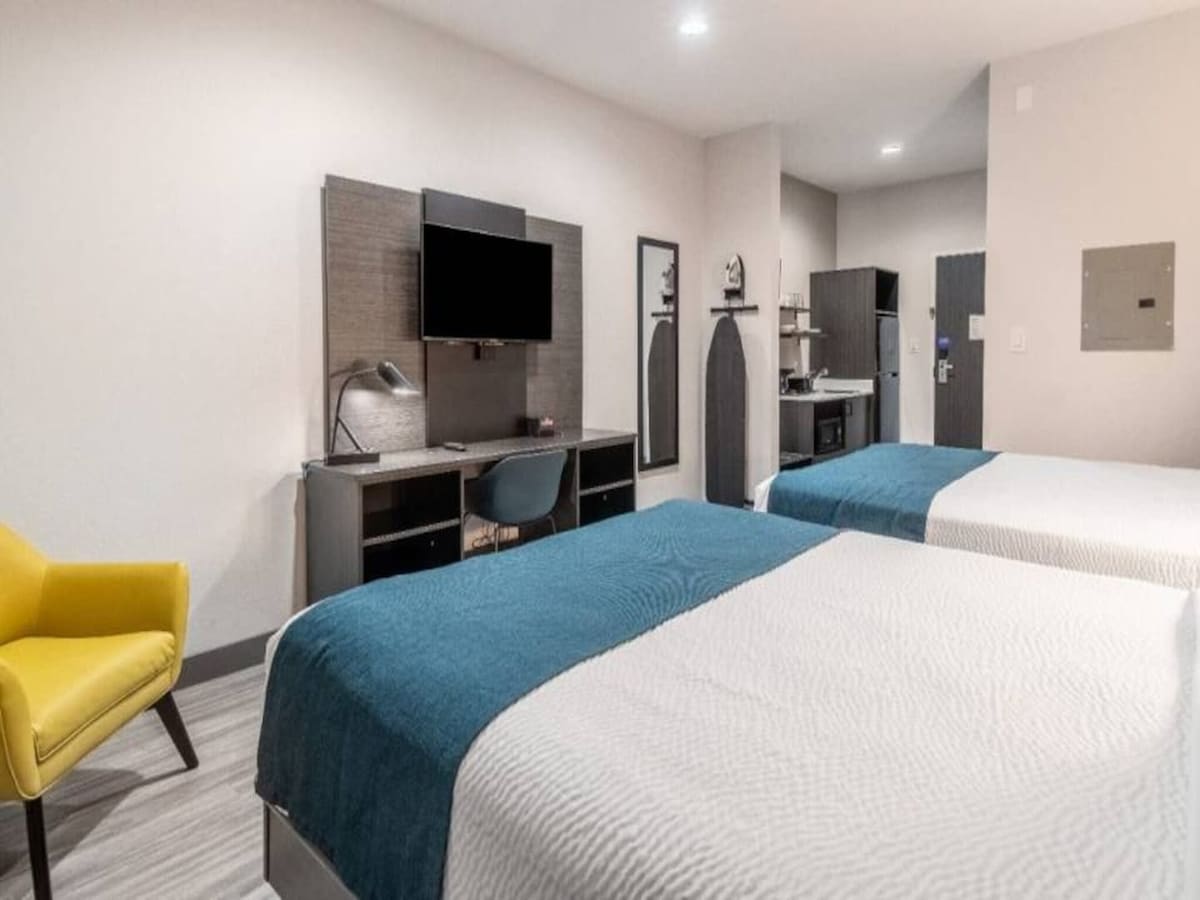 A spacious room features two queen beds dressed in blue and white linens. A work desk with a chair and lamp is positioned against the wall, next to a mounted television. A small kitchenette area is visible, including a refrigerator and microwave. A yellow accent chair adds a pop of color.