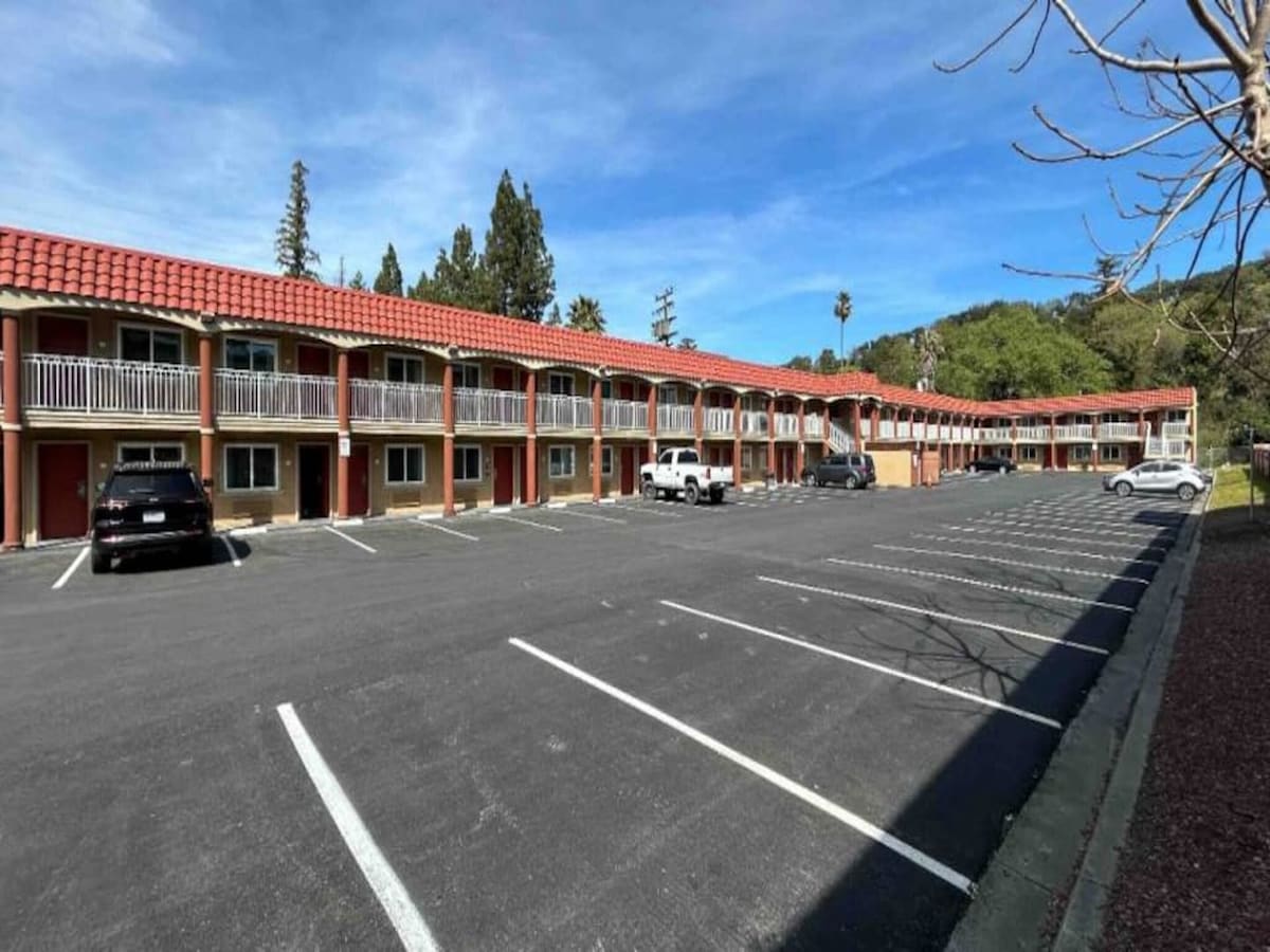 A view of the motel's exterior highlights a two-story structure with a red-tiled roof and multiple access points. Several parking spaces are visible in the foreground, accompanied by trees in the background and a clear blue sky overhead.